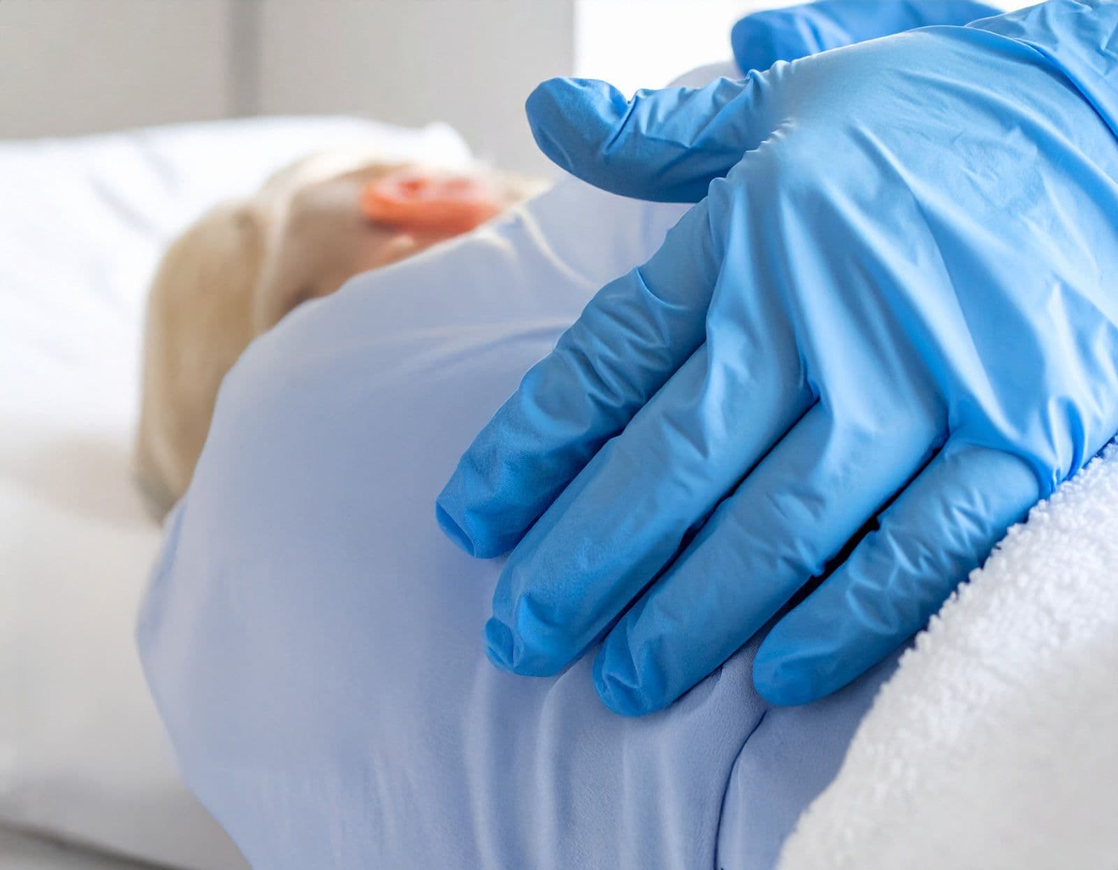 Ältere Person liegt seitlich im Bett, Hände mit blauen Handschuhen berühren behutsam den Rücken