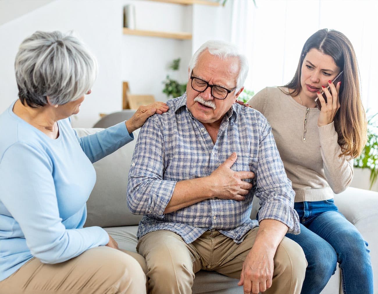 Senior sitzt auf Sofa und hält sich die Brust, zwei Frauen sitzen neben ihm, eine legt tröstend die Hand auf seine Schulter