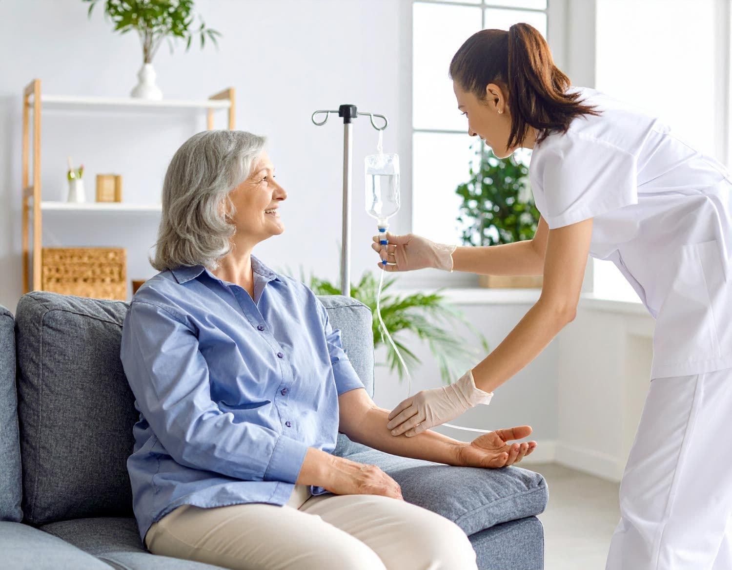 Ältere Frau sitzt entspannt auf einem Sofa, eine Pflegekraft hält eine Infusionsflasche und berührt ihren Arm