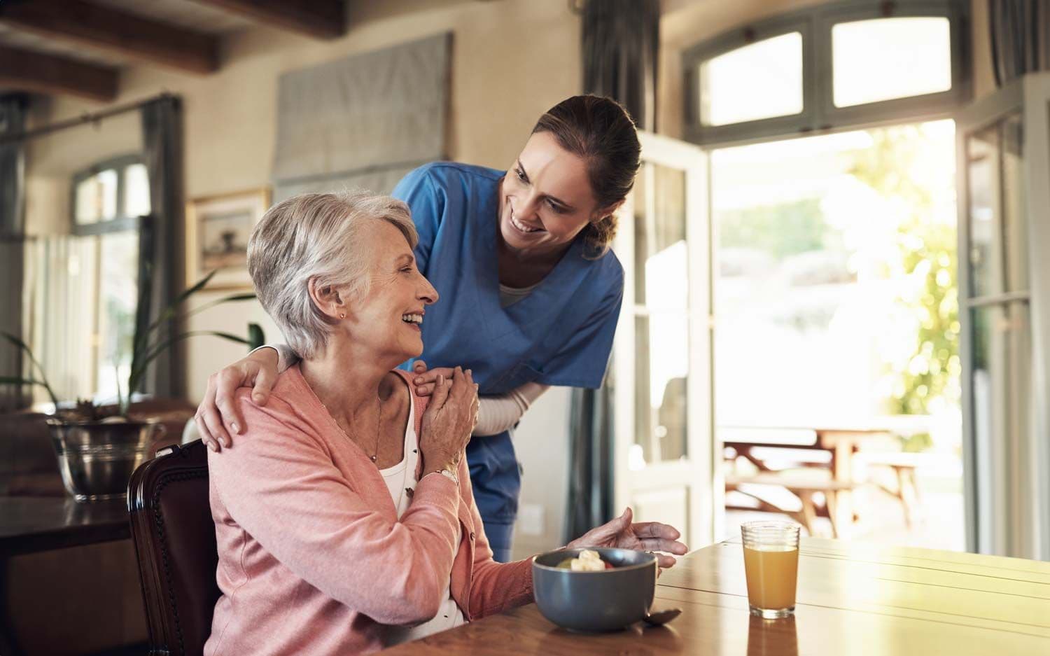 Pflegekraft berührt fürsorglich die Schultern einer älteren Frau, die am Tisch mit Frühstück sitzt