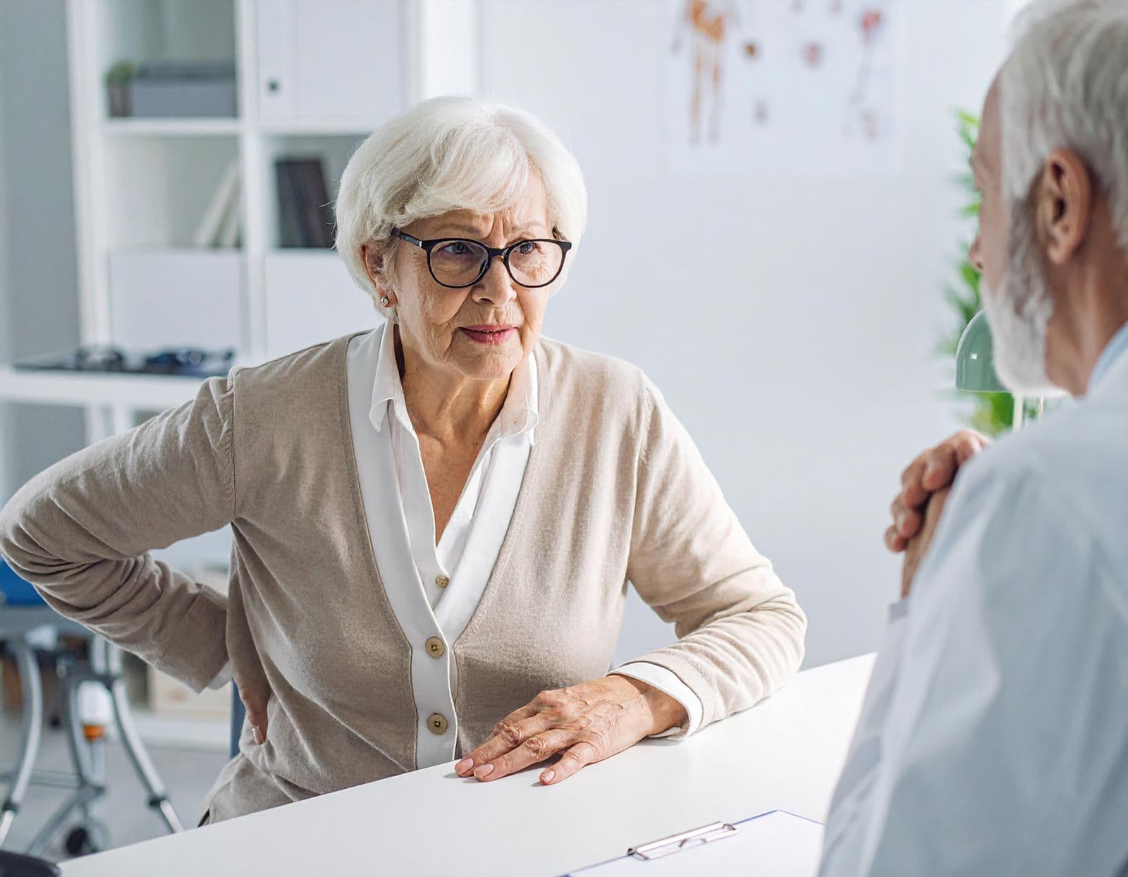 Ältere Frau mit Rückenschmerzen sitzt beim Arzt, der ihr zuhört