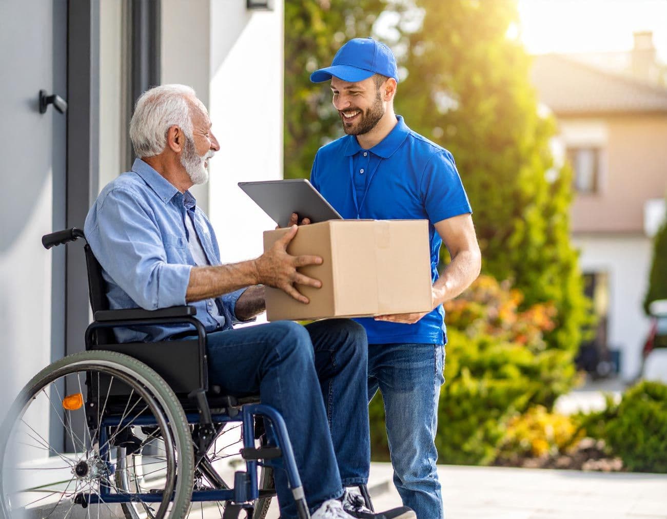 Älterer Mann im Rollstuhl erhält ein großes Paket von einem Lieferboten vor einem Haus
