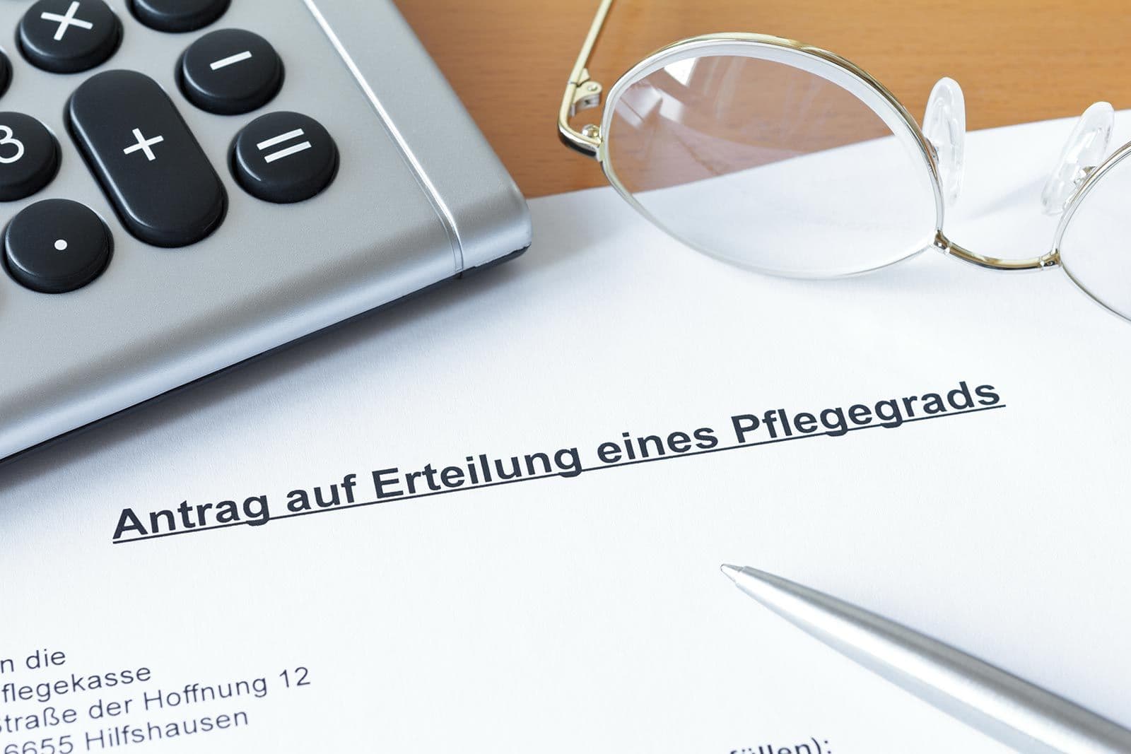 Antrag auf Erteilung eines Pflegegrads mit Taschenrechner, Brille und Kugelschreiber auf Holztisch
