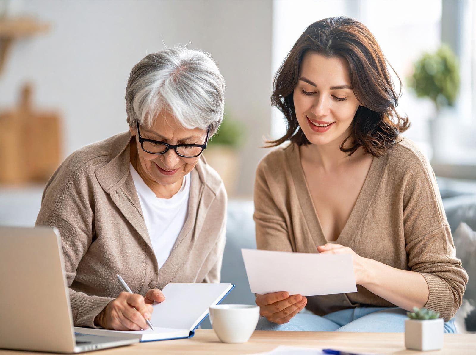 Junge Frau hilft älterer Frau, Dokumente auszufüllen, beide sitzen am Tisch mit Laptop und Unterlagen