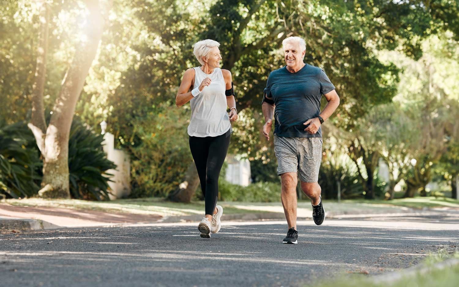 Älteres Paar joggt gemeinsam auf einer sonnigen Straße, umgeben von Bäumen