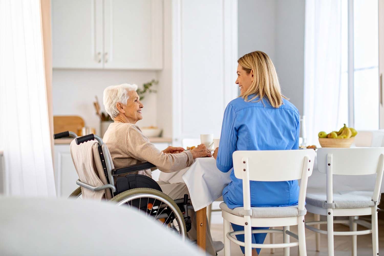 Eine Pflegekraft sitzt am Tisch mit einer älteren Frau im Rollstuhl, sie trinken gemeinsam Tee zu Hause
